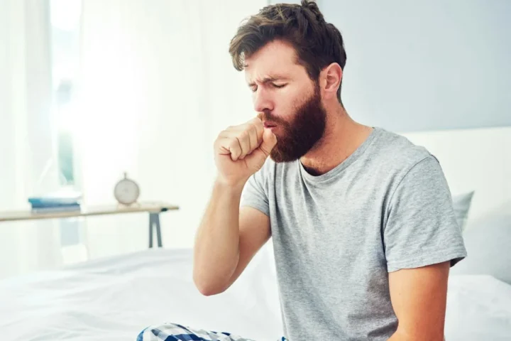 A man seated on a bed with his hand near his mouth while coughing.