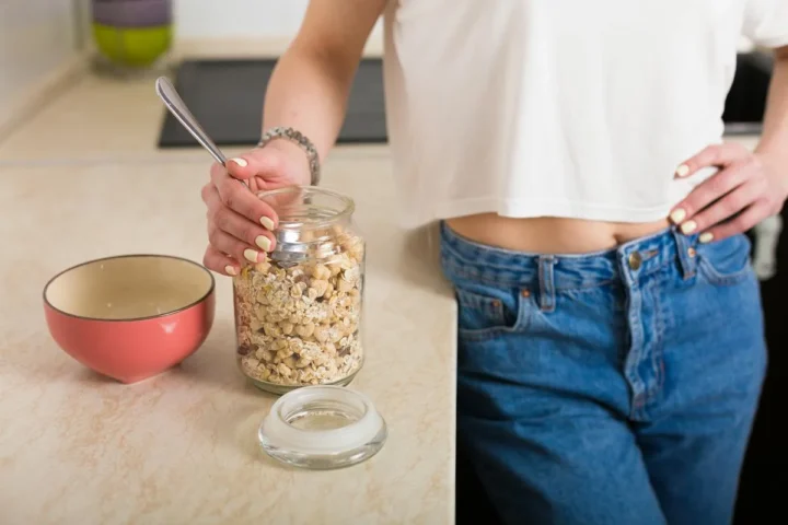 A person adding oats to a bowl beside a jar of rolled oats.