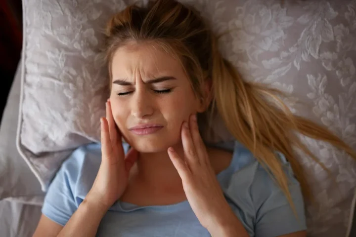 A woman in bed holding the side of her jaw.